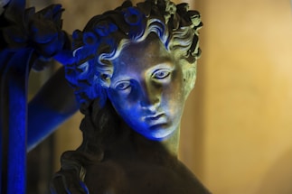 A close-up of a classical stone sculpture of a human face illuminated with dramatic blue lighting. The sculpture features intricate details with flowing hair and a serene expression, set against a softly blurred background.