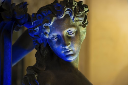 A close-up of a classical stone sculpture of a human face illuminated with dramatic blue lighting. The sculpture features intricate details with flowing hair and a serene expression, set against a softly blurred background.
