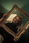 A person is smiling while holding an ornate, empty picture frame in front of them. The frame is golden with intricate detailing. The background is softly lit in green hues, and there is a lamp in the background providing warm light.