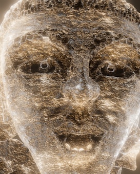 A close-up view of a textured stone sculpture of a face, illuminated to enhance its details. The sculpture's features include deeply set eyes, a prominent nose, and a slightly open mouth. The surface appears rugged with a metallic sheen, capturing shades of gold and silver.