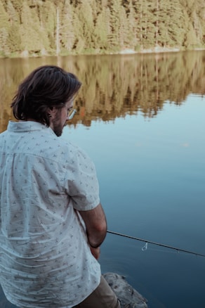A person with a patterned shirt and glasses is fishing by a calm lake. The water reflects a dense forest of green trees in the background. The atmosphere seems serene and peaceful.