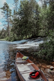 A wooden canoe is placed at the edge of a calm river, surrounded by dense greenery and tall trees. The river has a gentle waterfall in the background, creating a peaceful natural setting. Life jackets and paddles are visible inside the canoe.