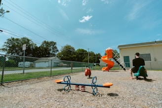A playground with a variety of equipment including a blue and orange seesaw, a red spring rider, and an orange spiral slide is seen. A man in a black shirt and cap is sitting on a green rocking toy shaped like an animal. The playground is enclosed by a chain-link fence, and there are trees and a building in the background under a clear blue sky.