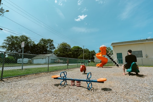 A playground with a variety of equipment including a blue and orange seesaw, a red spring rider, and an orange spiral slide is seen. A man in a black shirt and cap is sitting on a green rocking toy shaped like an animal. The playground is enclosed by a chain-link fence, and there are trees and a building in the background under a clear blue sky.