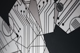 An abstract design featuring geometric shapes and circuit-like patterns with black lines on a light gray background. The intersecting lines and circles resemble a circuit board, creating a modern and technological aesthetic.