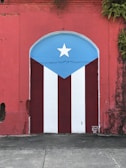 A vibrant mural painted on a wall features the Puerto Rican flag. The design includes vertical red and white stripes with a blue triangle on top, displaying a white star at its center. The surrounding wall is weathered red with ivy or plants growing from the top.