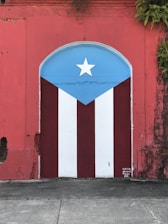 A vibrant mural painted on a wall features the Puerto Rican flag. The design includes vertical red and white stripes with a blue triangle on top, displaying a white star at its center. The surrounding wall is weathered red with ivy or plants growing from the top.
