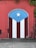 A vibrant mural painted on a wall features the Puerto Rican flag. The design includes vertical red and white stripes with a blue triangle on top, displaying a white star at its center. The surrounding wall is weathered red with ivy or plants growing from the top.