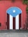A vibrant mural painted on a wall features the Puerto Rican flag. The design includes vertical red and white stripes with a blue triangle on top, displaying a white star at its center. The surrounding wall is weathered red with ivy or plants growing from the top.