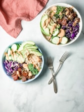 Two vibrant poke bowls filled with fresh ingredients including sliced avocado, cucumber, purple cabbage, and seaweed salad are arranged on a light surface. Each bowl is topped with a drizzle of creamy dressing. Two metallic forks lie on the surface, and a soft pink cloth is partially visible in the corner.
