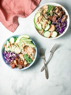 Two vibrant poke bowls filled with fresh ingredients including sliced avocado, cucumber, purple cabbage, and seaweed salad are arranged on a light surface. Each bowl is topped with a drizzle of creamy dressing. Two metallic forks lie on the surface, and a soft pink cloth is partially visible in the corner.
