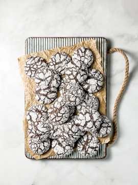 A tray with several chocolate crinkle cookies dusted with powdered sugar, placed on crinkled parchment paper. The cookies have a distinctive cracked surface pattern and are arranged on a ribbed metal tray with a rope handle.