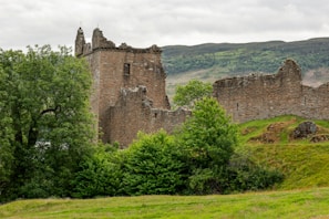 Ancient stone walls of a Scottish castle ruin surrounded by lush green hills under a cloudy sky.