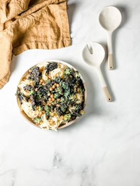 A plate of pasta topped with roasted chickpeas, kale, and broccoli. The dish is placed on a marble surface next to a pair of beige and white serving utensils. A crumpled brown cloth napkin is positioned to the upper left corner of the image.