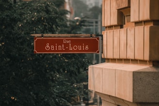 A street sign with 'Rue Saint-Louis' written on it is mounted on the corner of a building. The sign has a red background with white lettering and decorative edges. The building is constructed with large, light brown stones, and there are trees in the background.