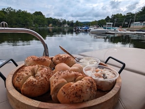 A wooden tray is topped with several sesame seed bagels alongside a couple of plastic containers filled with spreads or salads. In the background, there is a tranquil view of a lake with boats docked near the shoreline, surrounded by lush greenery and a cloudy sky.