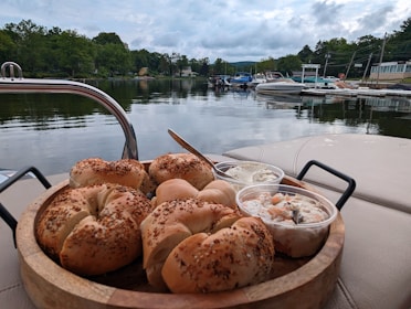 A wooden tray is topped with several sesame seed bagels alongside a couple of plastic containers filled with spreads or salads. In the background, there is a tranquil view of a lake with boats docked near the shoreline, surrounded by lush greenery and a cloudy sky.