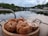 A wooden tray is topped with several sesame seed bagels alongside a couple of plastic containers filled with spreads or salads. In the background, there is a tranquil view of a lake with boats docked near the shoreline, surrounded by lush greenery and a cloudy sky.