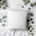 A plain, white pillow is surrounded by eucalyptus leaves arranged on a white fabric surface. The leaves and branches are symmetrically placed, creating a calming and soothing aesthetic.