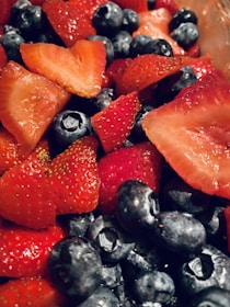Juicy red strawberries and plump blue blueberries are closely packed together, creating a vibrant display of fresh fruits. The strawberries are sliced and whole, showcasing their seeds and rich color, while the blueberries appear ripe and slightly glossy.