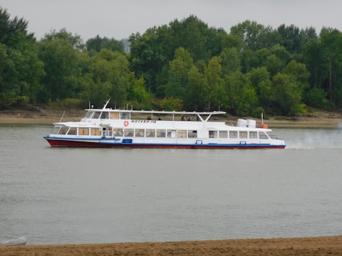 A white and blue riverboat with red accents is cruising along a calm river. The vessel has numerous windows and a prominent name written on its side. Dense, green forested banks form the background, and the sky appears overcast.