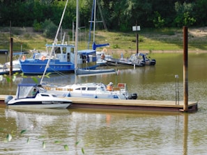 A calm riverside marina featuring several small boats docked by a wooden pier. The water is still with gentle ripples, and the area is surrounded by lush green foliage and small hills. One of the boats has a person in an orange life jacket seated at the back, indicating preparation for travel or maintenance.