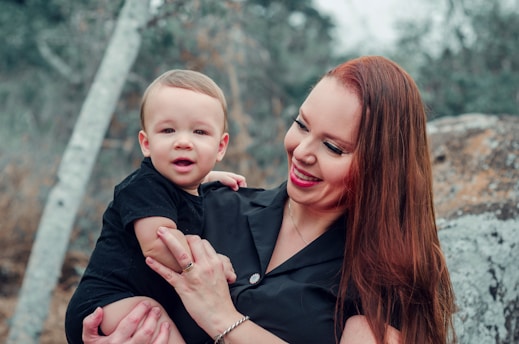 A woman with long, reddish-brown hair and a black outfit holds a young child in her arms. They are outdoors, surrounded by natural elements like rocks and trees. Both the woman and child appear to be smiling, conveying a sense of joy and affection.