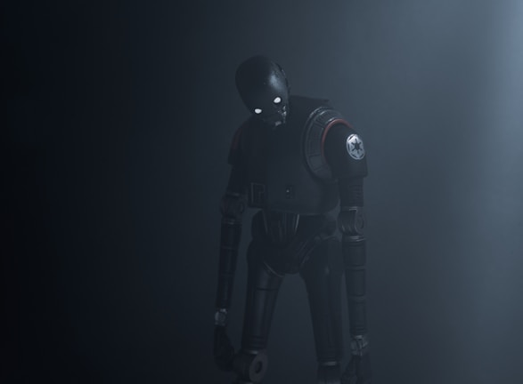 A humanoid robot with dark metallic body stands in a dimly lit environment, emitting an eerie presence. Its eyes glow white, providing a stark contrast to the dark surroundings. The robot's body is detailed with mechanical joints and a circular emblem on its shoulder.