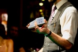A person wearing a formal outfit, including a vest and tie, holds a deck of cards with a blue design. The photo is taken in an indoor setting with dim lighting, and an audience can be seen blurred in the background, suggesting a performance or magic show.