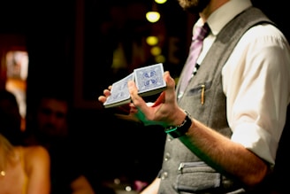 A person wearing a formal outfit, including a vest and tie, holds a deck of cards with a blue design. The photo is taken in an indoor setting with dim lighting, and an audience can be seen blurred in the background, suggesting a performance or magic show.