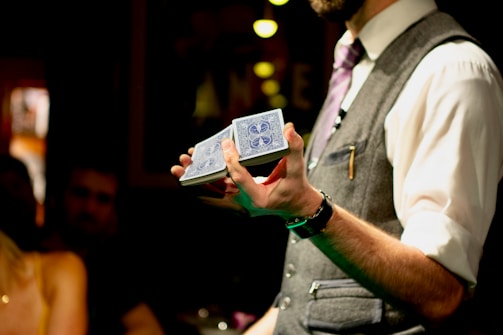 A person wearing a formal outfit, including a vest and tie, holds a deck of cards with a blue design. The photo is taken in an indoor setting with dim lighting, and an audience can be seen blurred in the background, suggesting a performance or magic show.