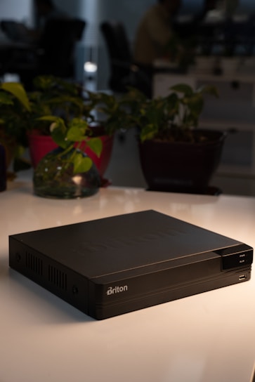 A black electronic device with the brand name 'Briton' is prominently placed on a white surface. In the background, there are various potted plants and blurred figures that suggest an office environment.