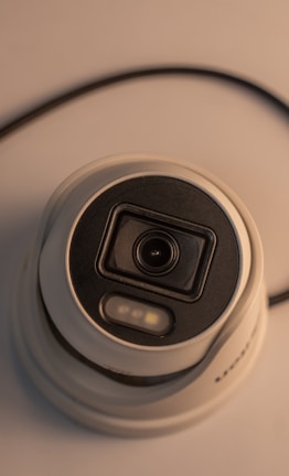 A close-up view of a dome-shaped security camera featuring a black lens housing and a white casing. The perspective highlights the circular design and electronic components, with a visible cable extending from the device.