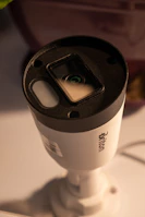A cylindrical security camera with the brand name visible on its side, positioned on a white surface. The camera lens and sensor are prominent, and it is attached to a mounting bracket.