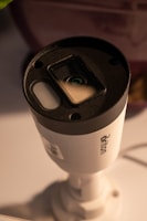 A cylindrical security camera with the brand name visible on its side, positioned on a white surface. The camera lens and sensor are prominent, and it is attached to a mounting bracket.