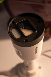 A cylindrical security camera with the brand name visible on its side, positioned on a white surface. The camera lens and sensor are prominent, and it is attached to a mounting bracket.