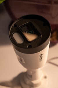 A cylindrical security camera with the brand name visible on its side, positioned on a white surface. The camera lens and sensor are prominent, and it is attached to a mounting bracket.
