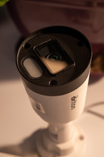 A cylindrical security camera with the brand name visible on its side, positioned on a white surface. The camera lens and sensor are prominent, and it is attached to a mounting bracket.