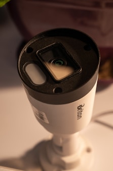 A cylindrical security camera with the brand name visible on its side, positioned on a white surface. The camera lens and sensor are prominent, and it is attached to a mounting bracket.