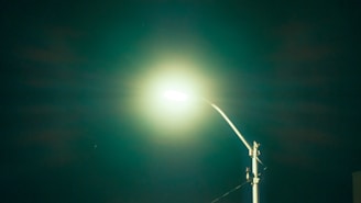 A bright streetlight stands illuminated against a dark night sky, casting a soft halo of light. The pole is metallic and slightly angled, with electrical components visible near the top.