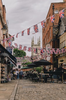 A charming cobblestone street lined with shops and outdoor seating, adorned with Union Jack flags. People are gathered, engaging in conversations and enjoying the atmosphere. The scene is set against the backdrop of a historical building with two large towers.