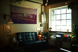 A cozy living room or lounge area with a dark leather sofa, decorated with string lights and various art pieces on the walls. A television displaying a color test pattern is placed on a retro-style cabinet. The room is warmly lit with small lamps and natural light from a large window adorned with plants and small figurines.