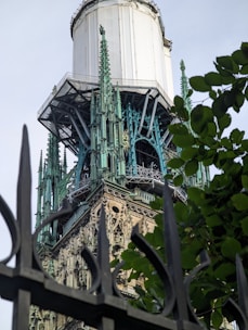 An ornate Gothic architectural structure with intricate detailing and greenish patina stands behind a dark wrought iron fence and leafy greenery. The structure appears to be under renovation, with a white covering partially obscuring the top portions.