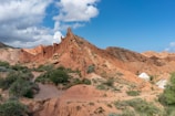 Rocky, reddish landscape featuring jagged formations under a blue sky with scattered clouds. Sparse vegetation consisting of green shrubs is present throughout the terrain. A few white yurts, traditional tent-like shelters, are scattered along the base of the hills.