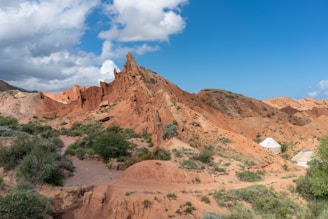 Rocky, reddish landscape featuring jagged formations under a blue sky with scattered clouds. Sparse vegetation consisting of green shrubs is present throughout the terrain. A few white yurts, traditional tent-like shelters, are scattered along the base of the hills.