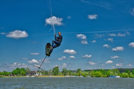 A person engaged in kite foiling, suspended in mid-air with a board and kite. The background features a serene lake, surrounded by lush greenery and a clear blue sky dotted with fluffy clouds.
