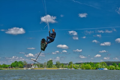 A person engaged in kite foiling, suspended in mid-air with a board and kite. The background features a serene lake, surrounded by lush greenery and a clear blue sky dotted with fluffy clouds.
