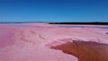A vast, pink-hued salt lake under a clear blue sky spans the foreground, showcasing sweeping natural formations and color gradients. The smooth surface features a darker patch of water that contrasts with the lighter pink areas, while the horizon is defined by a thin strip of dark land.