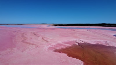 A vast, pink-hued salt lake under a clear blue sky spans the foreground, showcasing sweeping natural formations and color gradients. The smooth surface features a darker patch of water that contrasts with the lighter pink areas, while the horizon is defined by a thin strip of dark land.