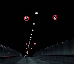 A dark tunnel interior with illuminated speed limit signs indicating 80 km/h. The tunnel is lined with ceiling lights running down the center, and a single vehicle is visible in the distance moving away.